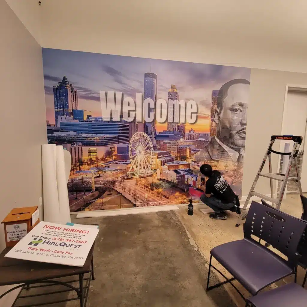 Person applying a large mural with a city skyline and "Welcome" text on a wall.