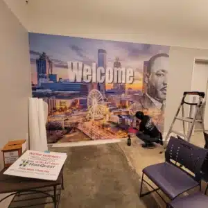 Person applying a large mural with a city skyline and "Welcome" text on a wall.