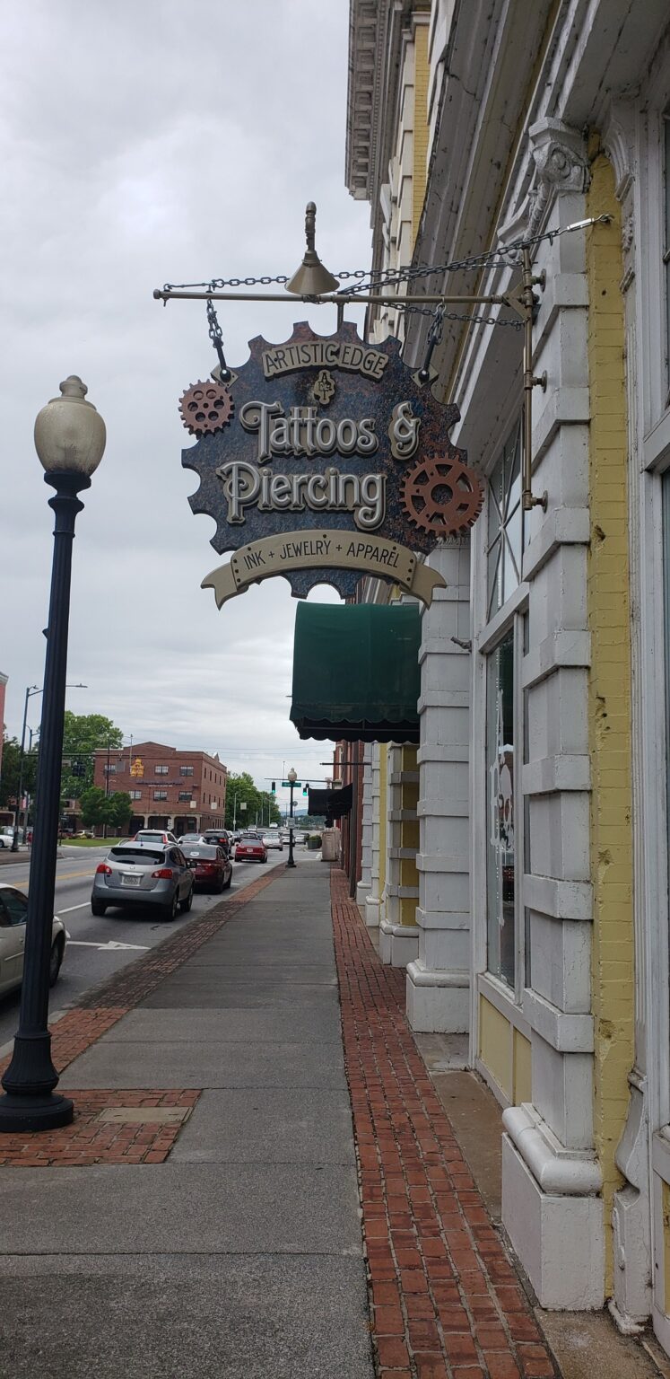 Steampunk gear-shaped hanging blade sign on ornate bracket, Artistic Edge Tattoos & Piercing, by High Stakes Creative