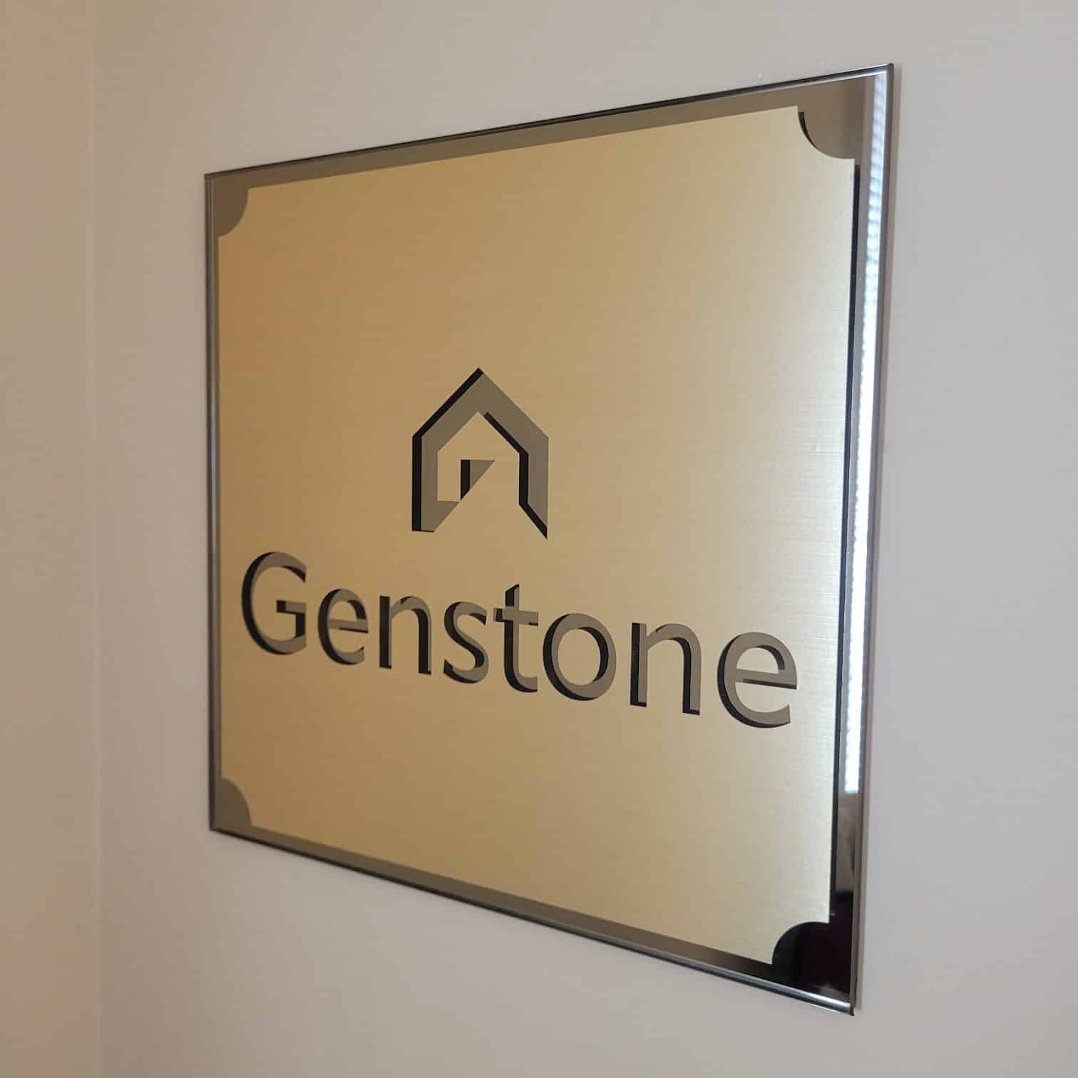 Brushed-gold logo plaque with dark border and corner accents on white wall, Genstone, by High Stakes Creative